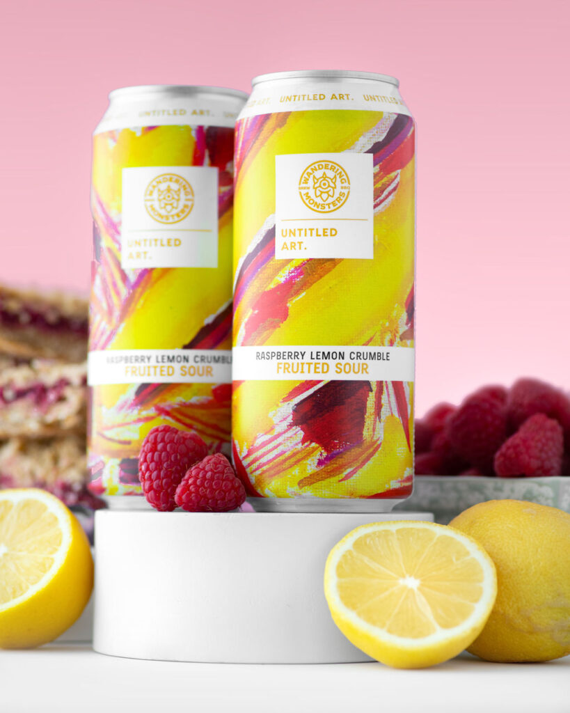 Two cans of raspberry lemon crumble fruited sour beer displayed with fresh raspberries and lemon slices against a pink background.
