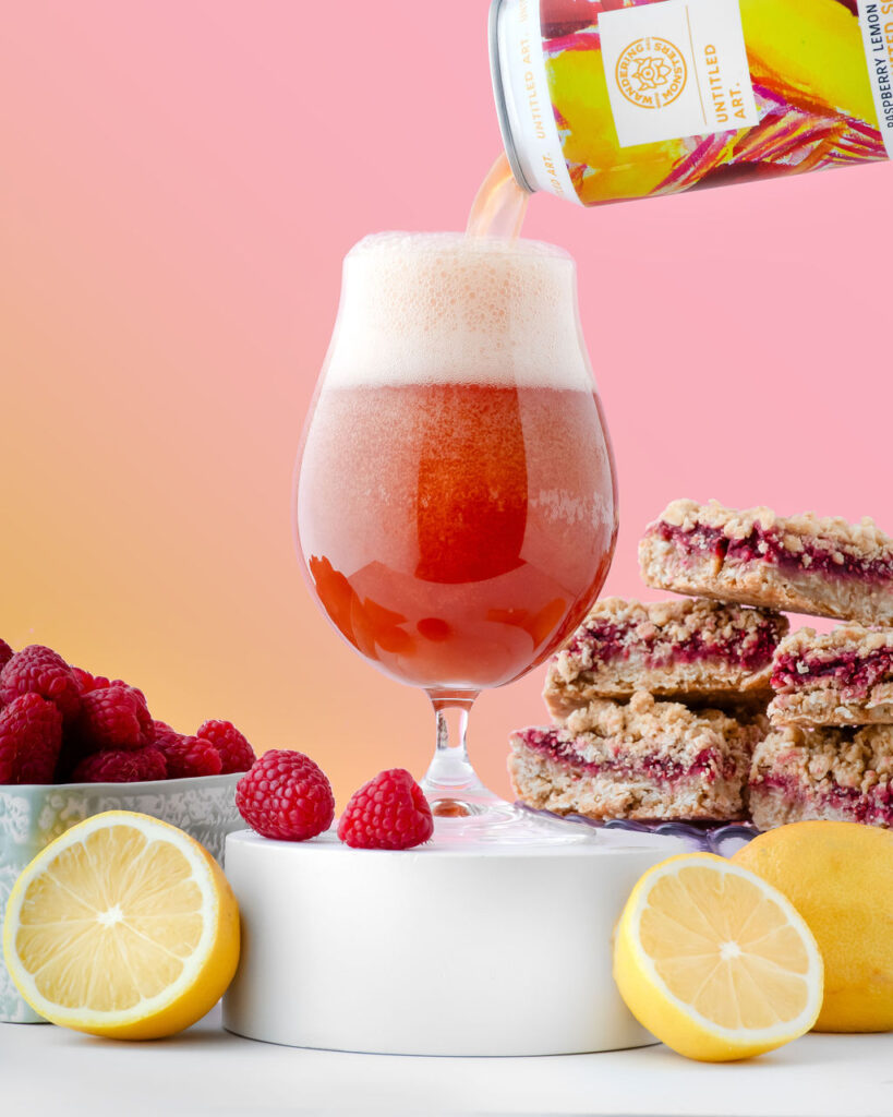 Golden ale being poured into a glass beside raspberries, lemon slices, and raspberry pastries on a vibrant background.