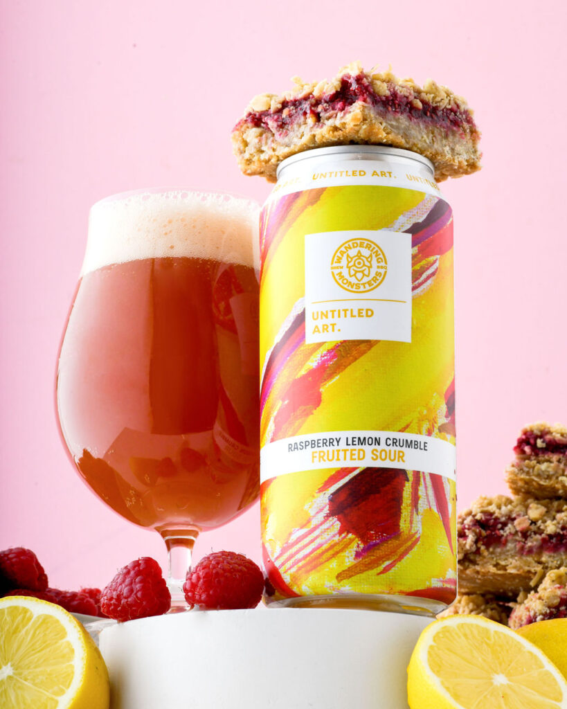 A can of "raspberry lemon crumble fruited sour" beer by untitled art, placed next to a glass of the poured beer, garnished with raspberries, lemon slices, and crumble bars against a pink background.