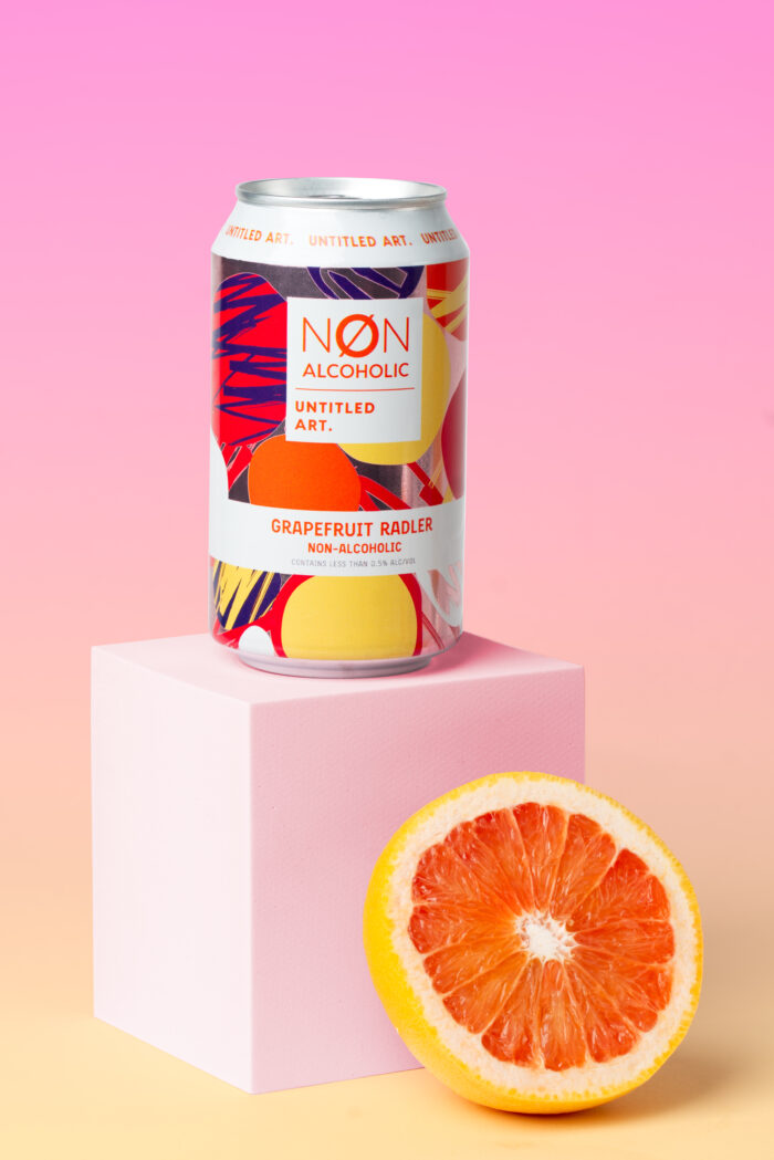 A can from a six-pack of Non-Alcoholic Grapefruit Radler is placed on a pink cube with half a grapefruit next to it, set against a pink gradient background.